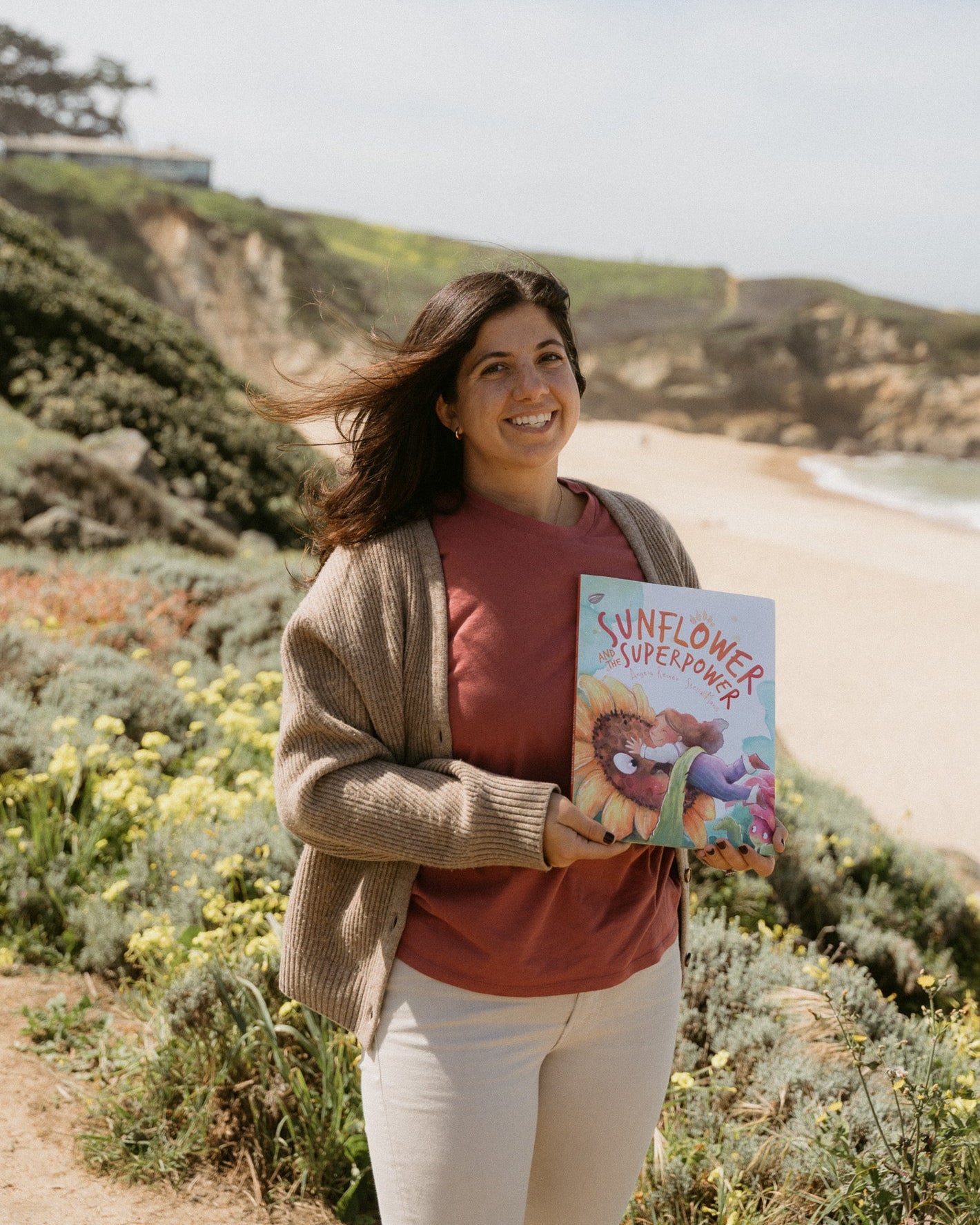 Woman holding a book titled 'Sunflower Superpower' on a beach with cliffs and greenery in the background.