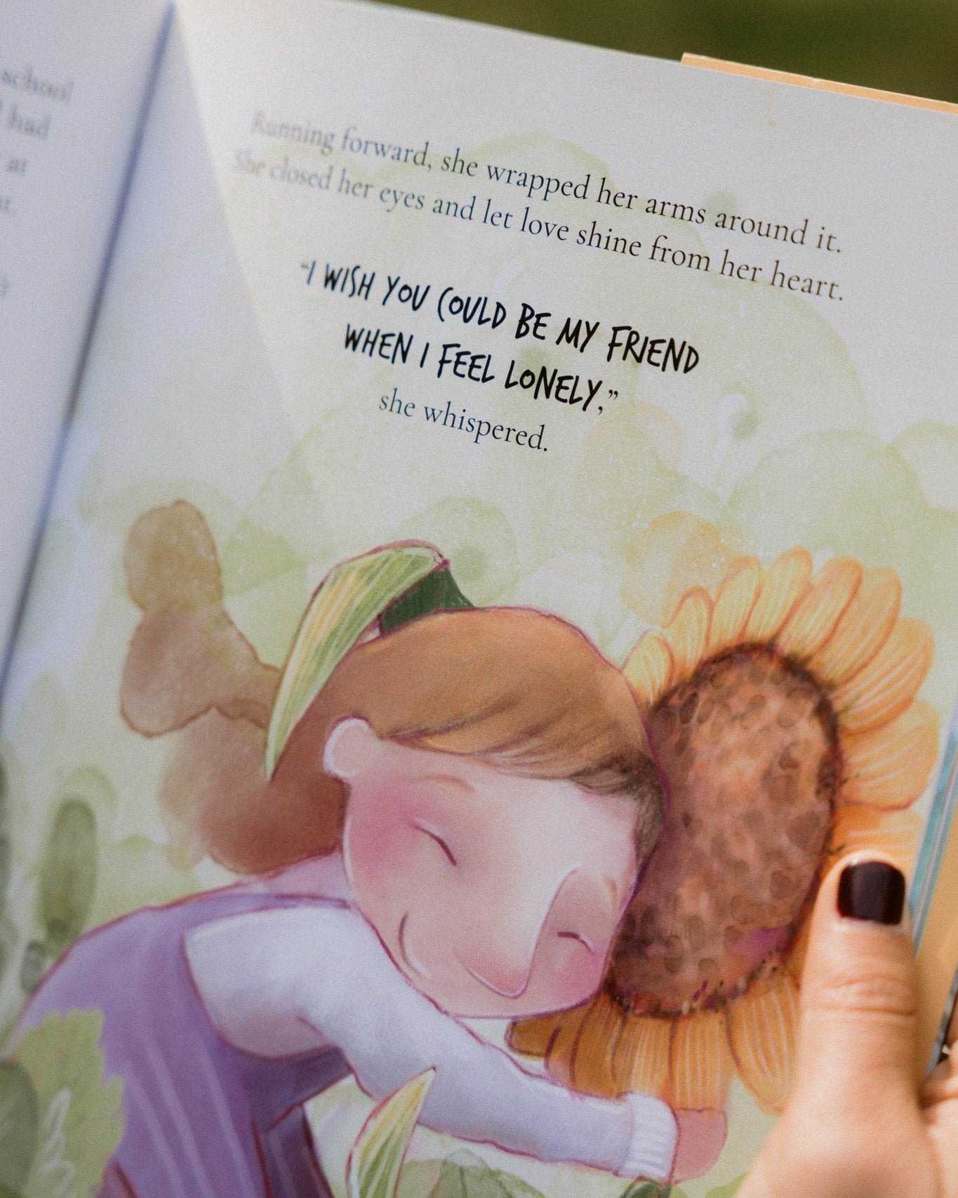 Illustrated book page with a girl hugging a sunflower and text, held by a hand.